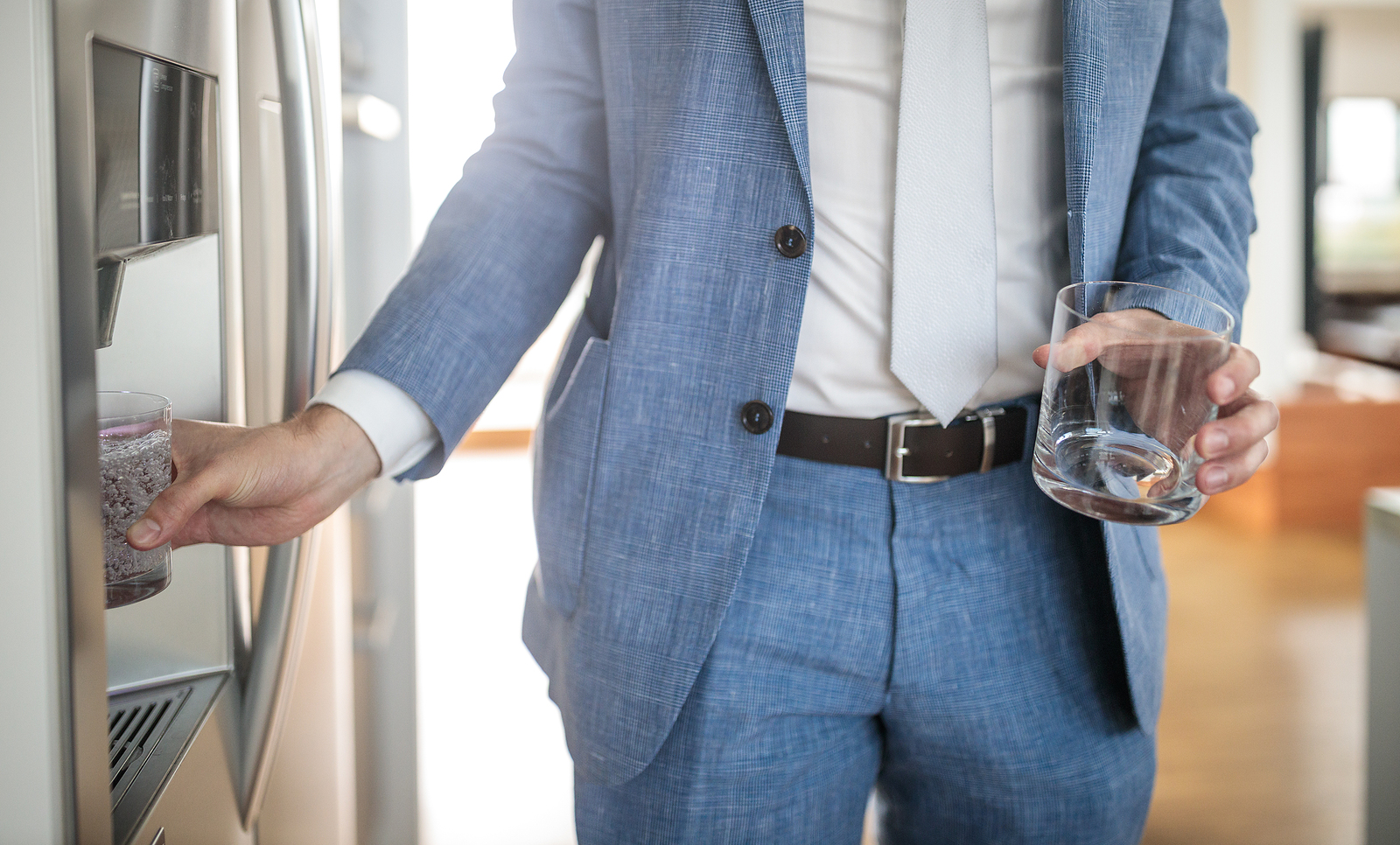 A man in a blue suit holding a glass of water from a fridge dispenser in a modern kitchen.