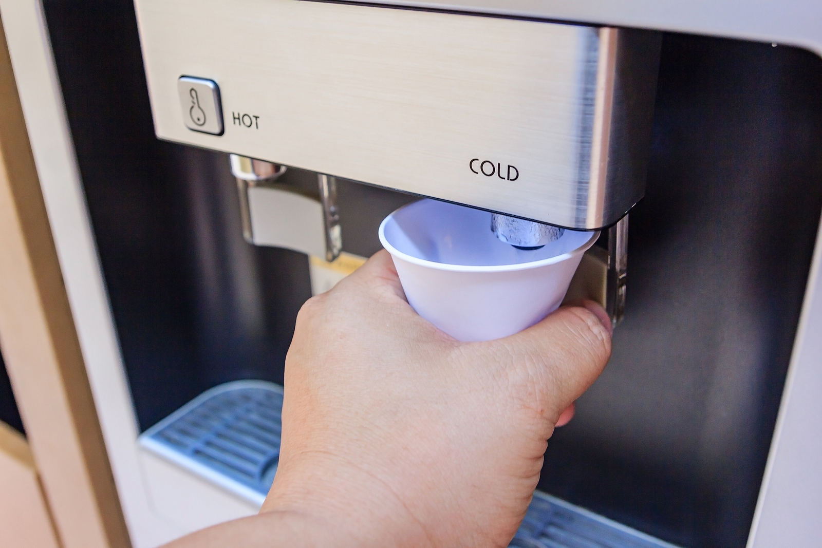 A hand holding a paper cup under a water cooler dispenser with hot and cold water options.