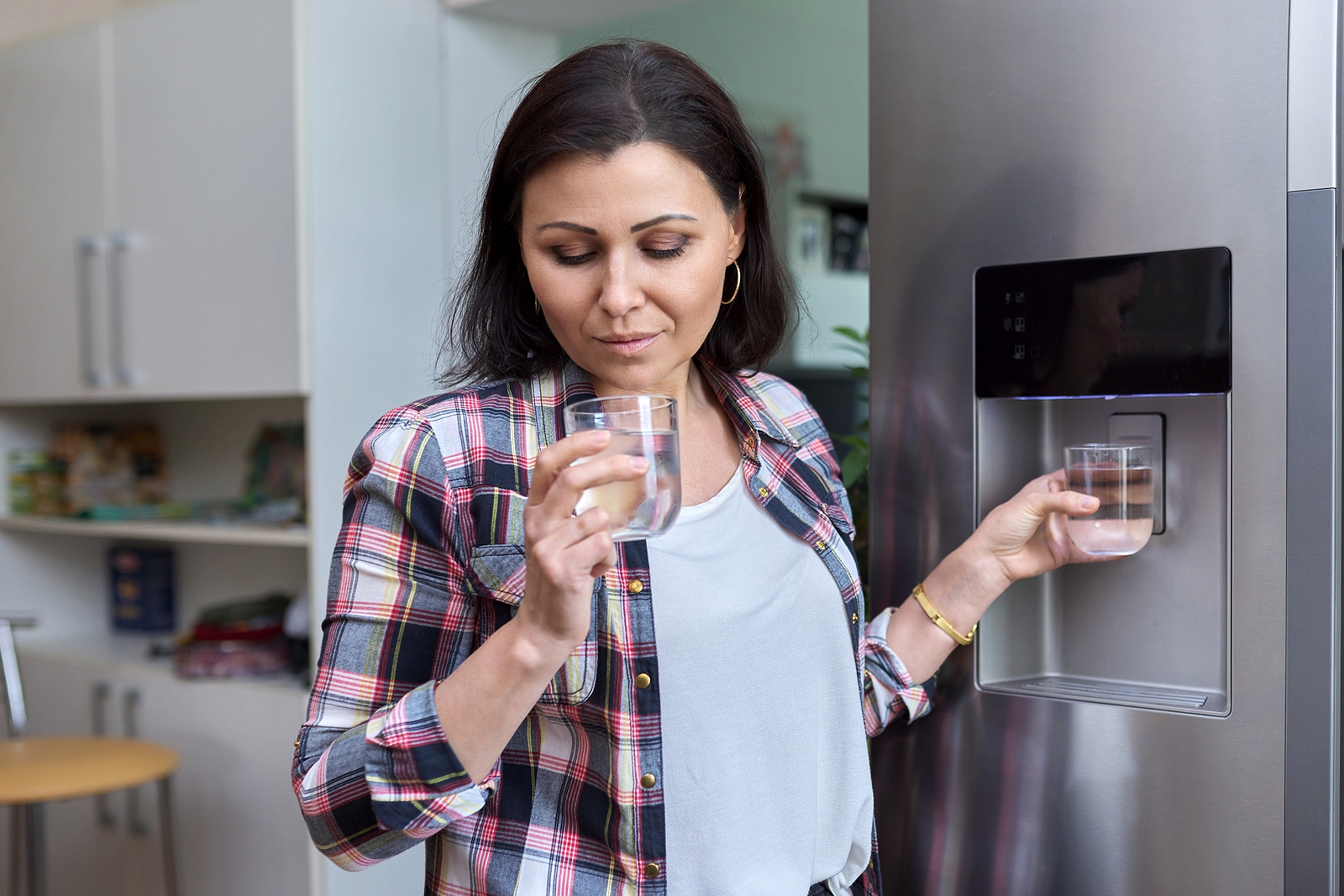 A woman in a plaid shirt holding two glasses of water from a fridge dispenser in a home kitchen.