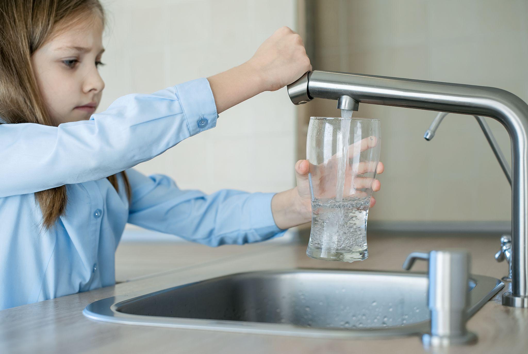 A child in a blue shirt filling a glass with water from a kitchen faucet, standing over a sink.