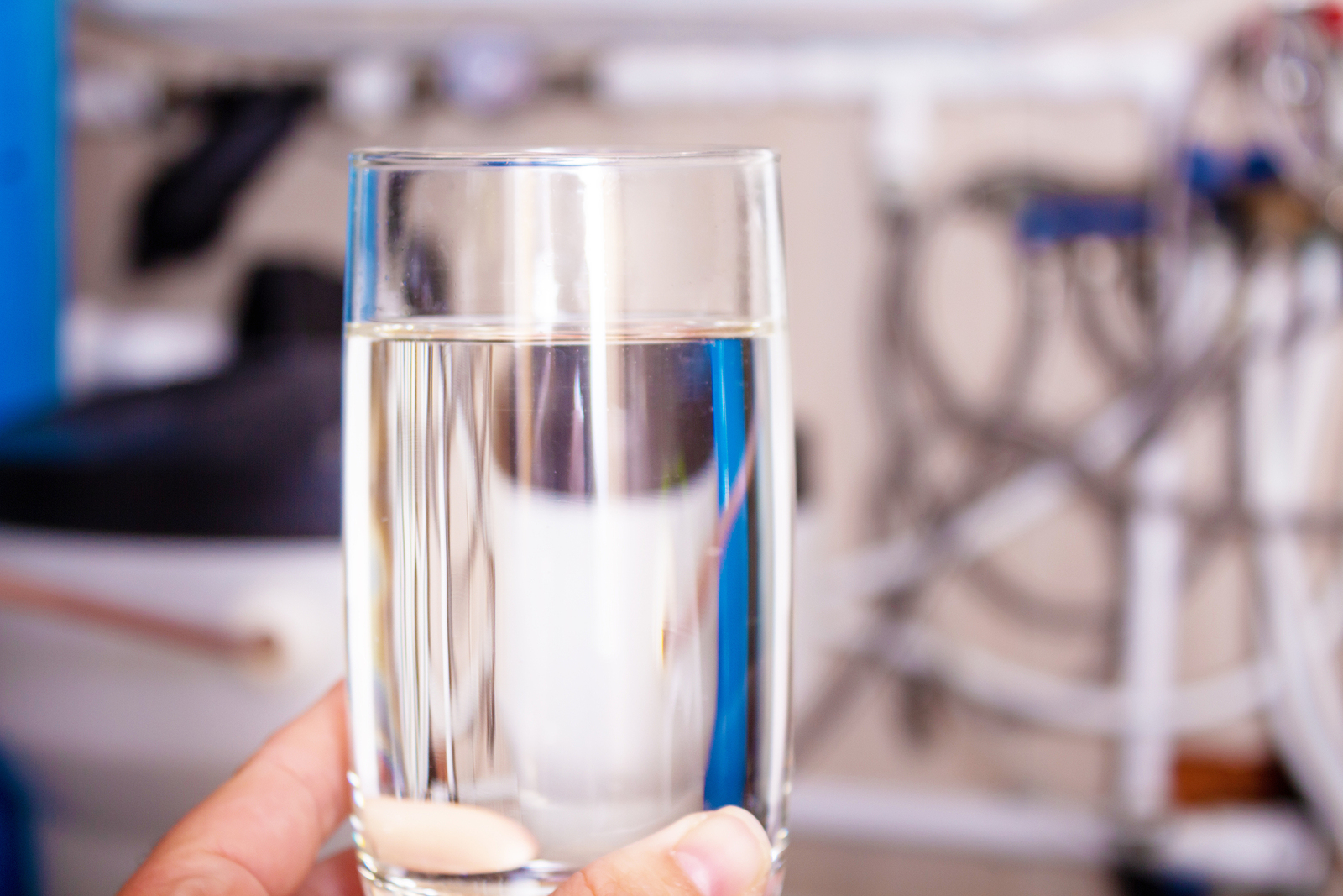 A hand holding a glass of clear water, set against a blurred background of a water softener system