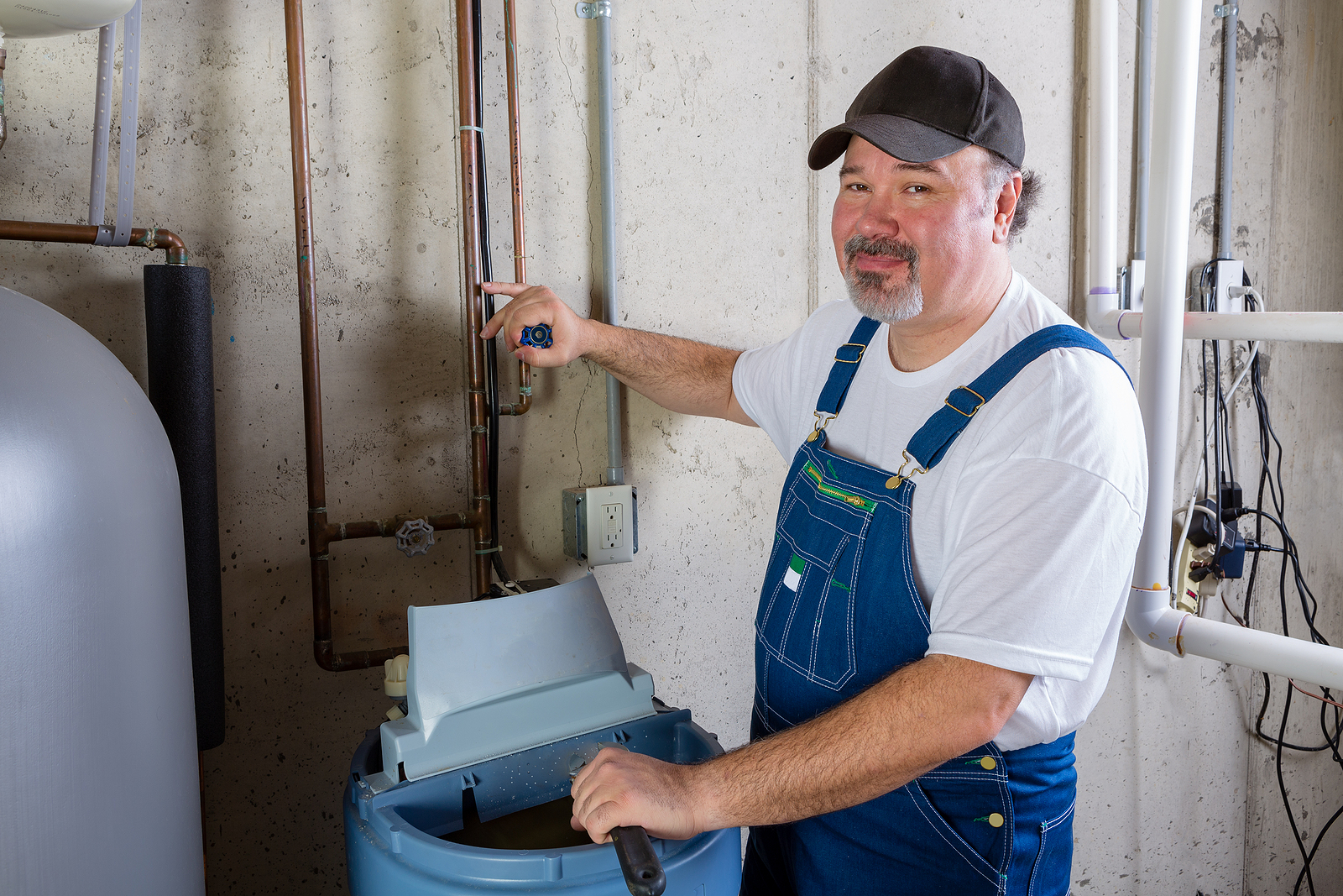 A technician in blue overalls adjusting a water softener tank in a basement, with pipes and tools in the background.