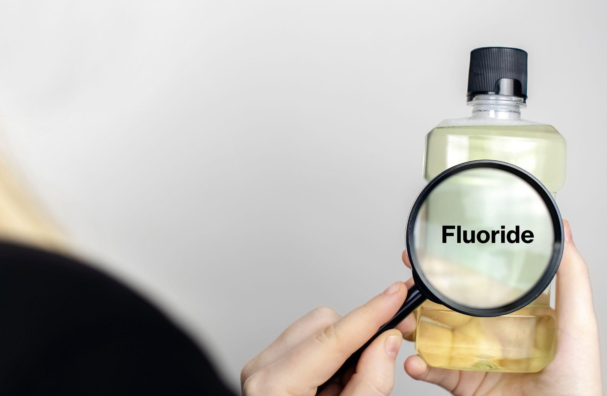 Person holding a bottle labeled fluoride, using a magnifying glass to highlight fluoride content in drinking water.