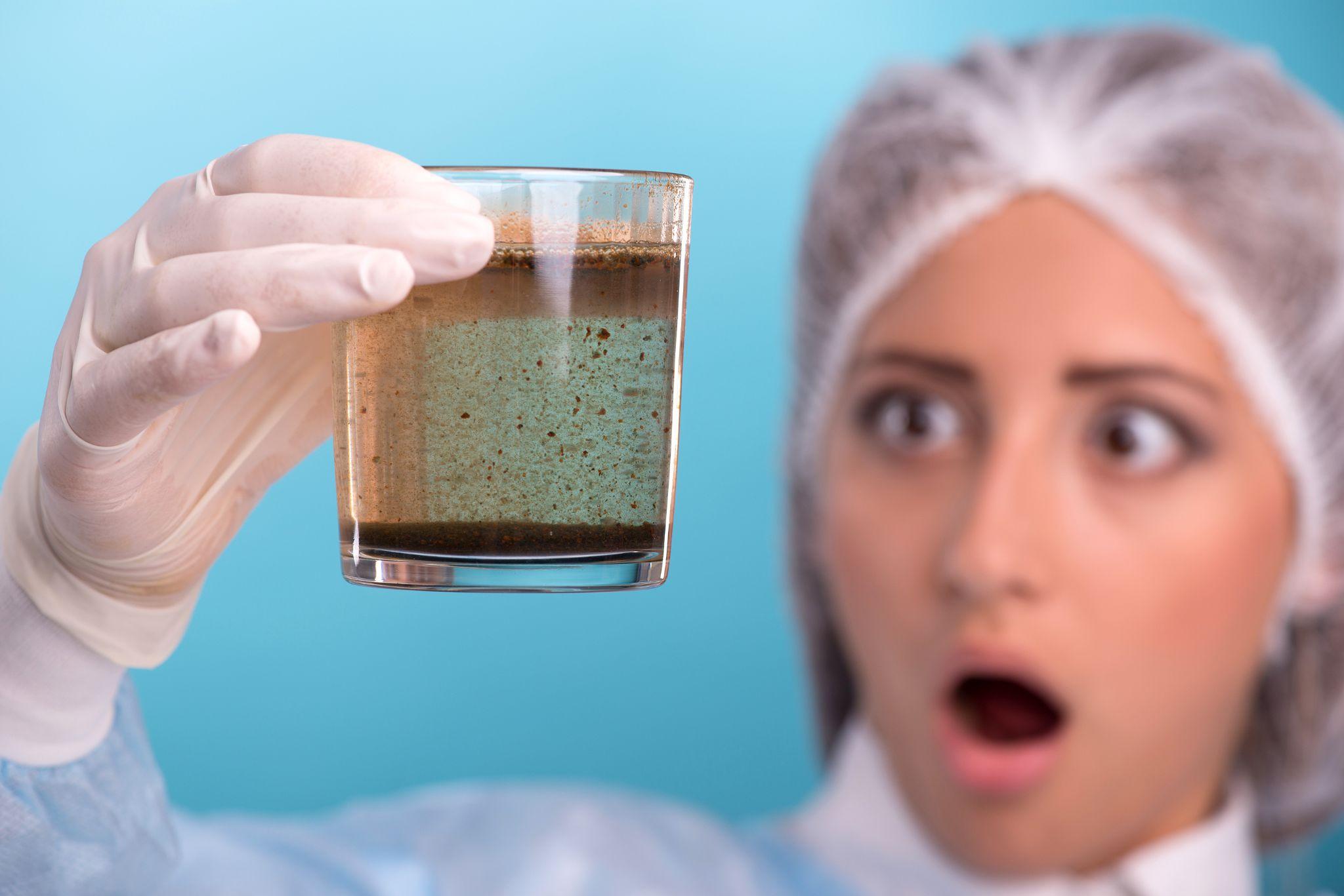 Lab technician holding a glass of visibly dirty water with a shocked expression, highlighting tap water contamination concerns in San Diego